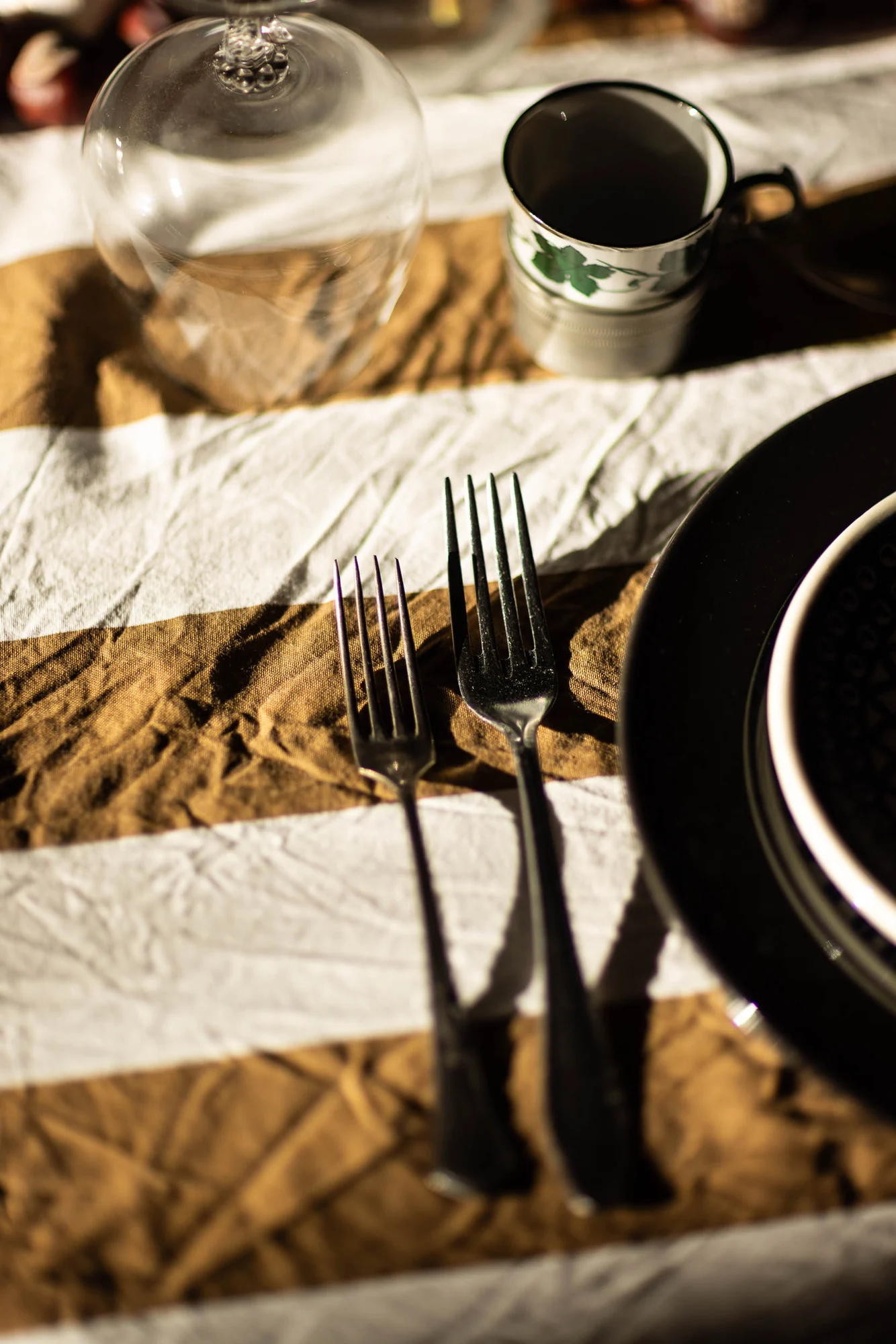 Gedeck aus der Nähe: gestreifter Leinenläufer in Ocker und Weiß, umgedrehtes Weinglas, Tasse mit grünem Weinlaubdekor, zwei alte Silbergabeln, schwarzer Teller