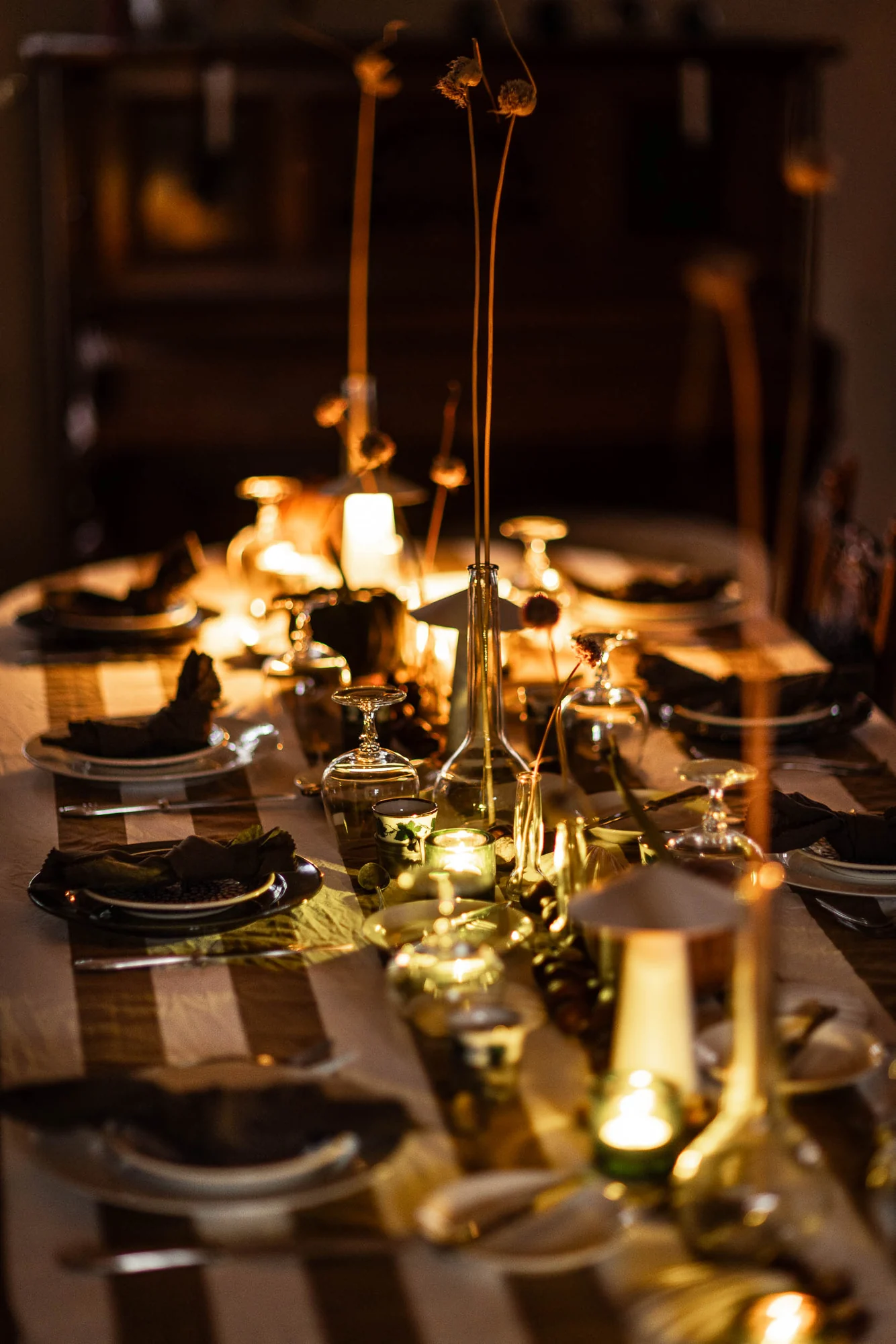 Die lange Tafel von der Stirnseite im Kerzenlicht, umgedrehte Gläser, dunkle Servietten und getrocknete Allium-Dolden in einer grünen Flasche